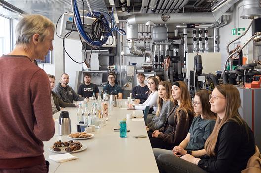 A group of students is sitting at a large table with snacks and drinks on it. In the foreground, a lecturer is speaking to them.